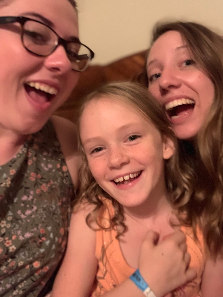 This picture is a selfie of three girls. The one on the left is wearing glasses and a green shirt with little yellow flowers. The one in the middle is younger and wearing a pink shirt. The one on the right has long curly dirty blonde hair. They are all smiling.