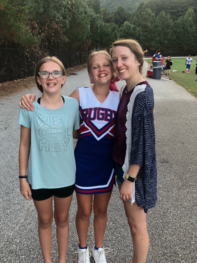 This picture is of three girls. The one on the left has her hair pulled back, a teal shirt, black shorts, and has glasses on. The middle girl is wearing a cheer uniform that is blue, red, and white. She is also wearing white tennis shoes and has her hair pulled back. The girl on the right is older and wearing a blue and white cardigan, maroon shirt, and jean shorts. Her hair is also pulled back. All three girls are smiling. This was taken on an asphalt sidewalk on a football field.