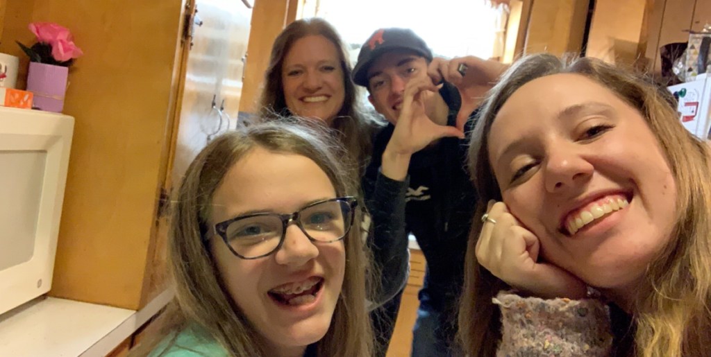 This picture is a selfie of three females and one male. There are two females, a younger girl and an older one, who are closest to the camera. They are both smiling. In the back there is an older women, who is smiling, and a young man, who is holding up his hands to make a heart.